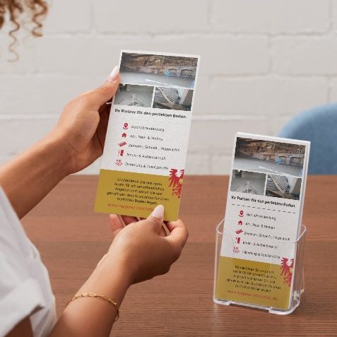 Zwei Informationsflyer in einer Halterung, gehalten von einer Person, auf einem Tisch.