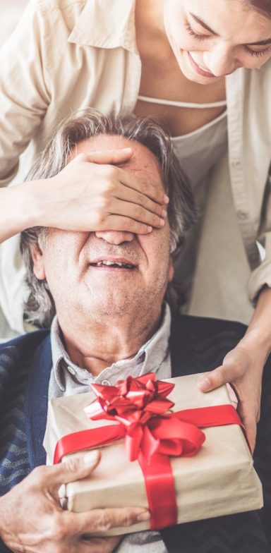 Einen älteren Mann werden von einer Frau die Augen zugehalten und es wird Ihm ein Geschenk überreicht